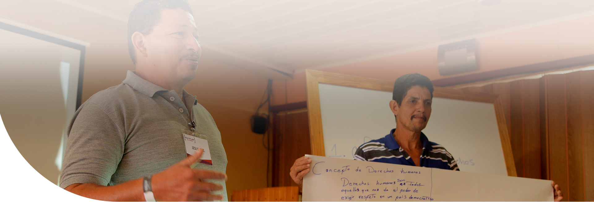 Dos hombres de pie exponiendo, uno de ellos sostiene un cartel